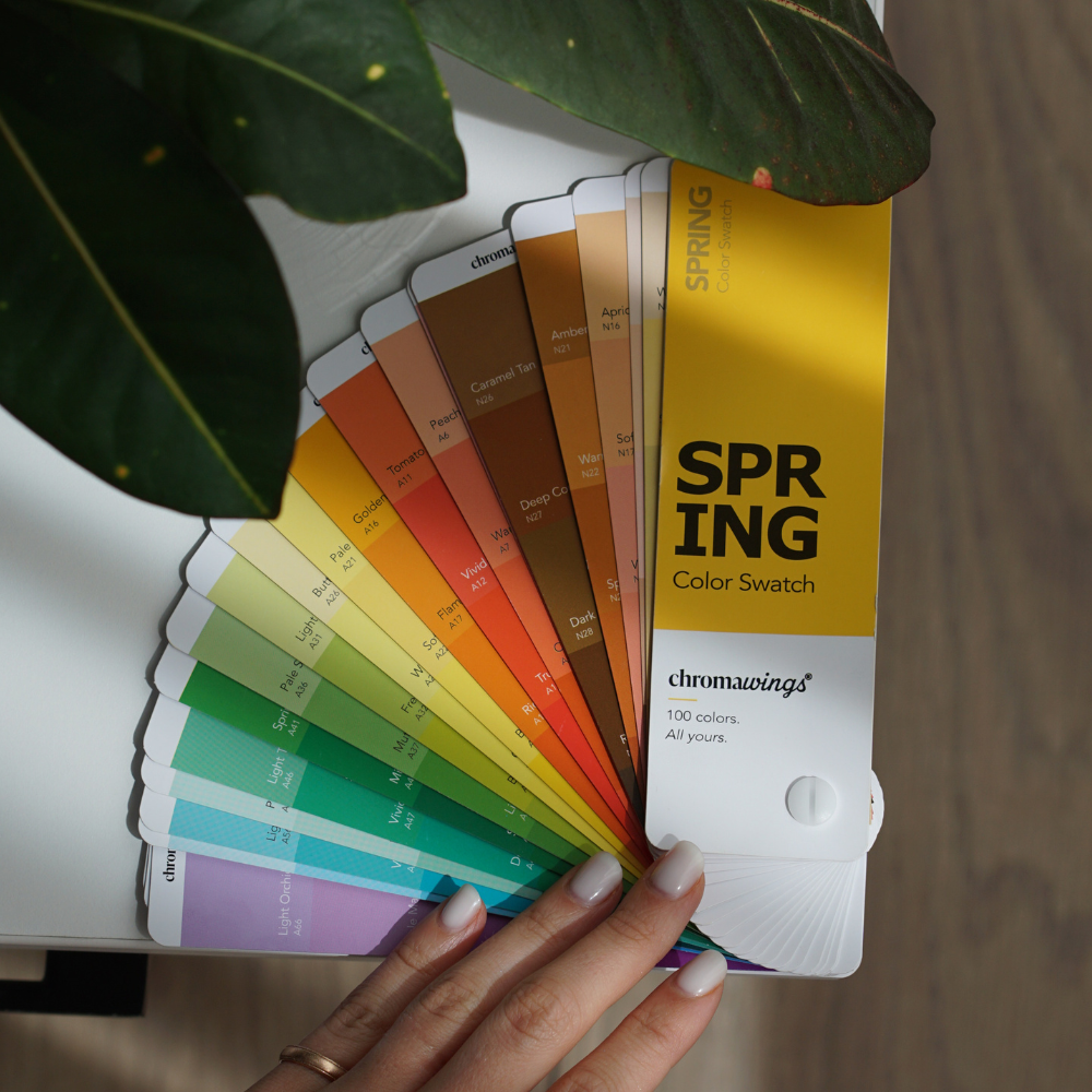 A hand holds a fanned-out Chromwings color swatch card set labeled 'SPRING Color Swatch,' showing multiple pastel and bright color options, with large green leaves partially covering the top of the fan on a light surface. #season_spring